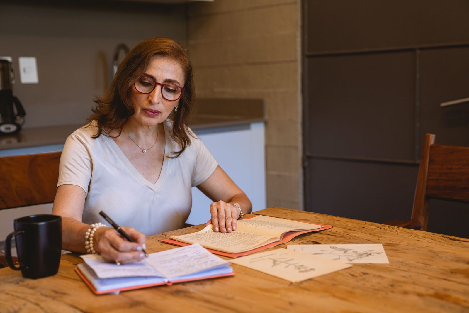 Eine Frau in ihren Fünfzigern schreibt an einem Küchentisch in ein Notizbuch, neben ihr steht eine Tasse